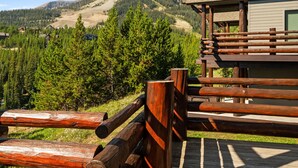 A wooden deck with a mountain view, a cabin with a sloped roof, and a forested area in the background.