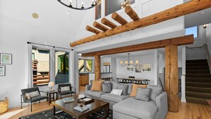 A spacious living room with a grey sectional sofa, wooden beams, and a staircase leading to an upper level.