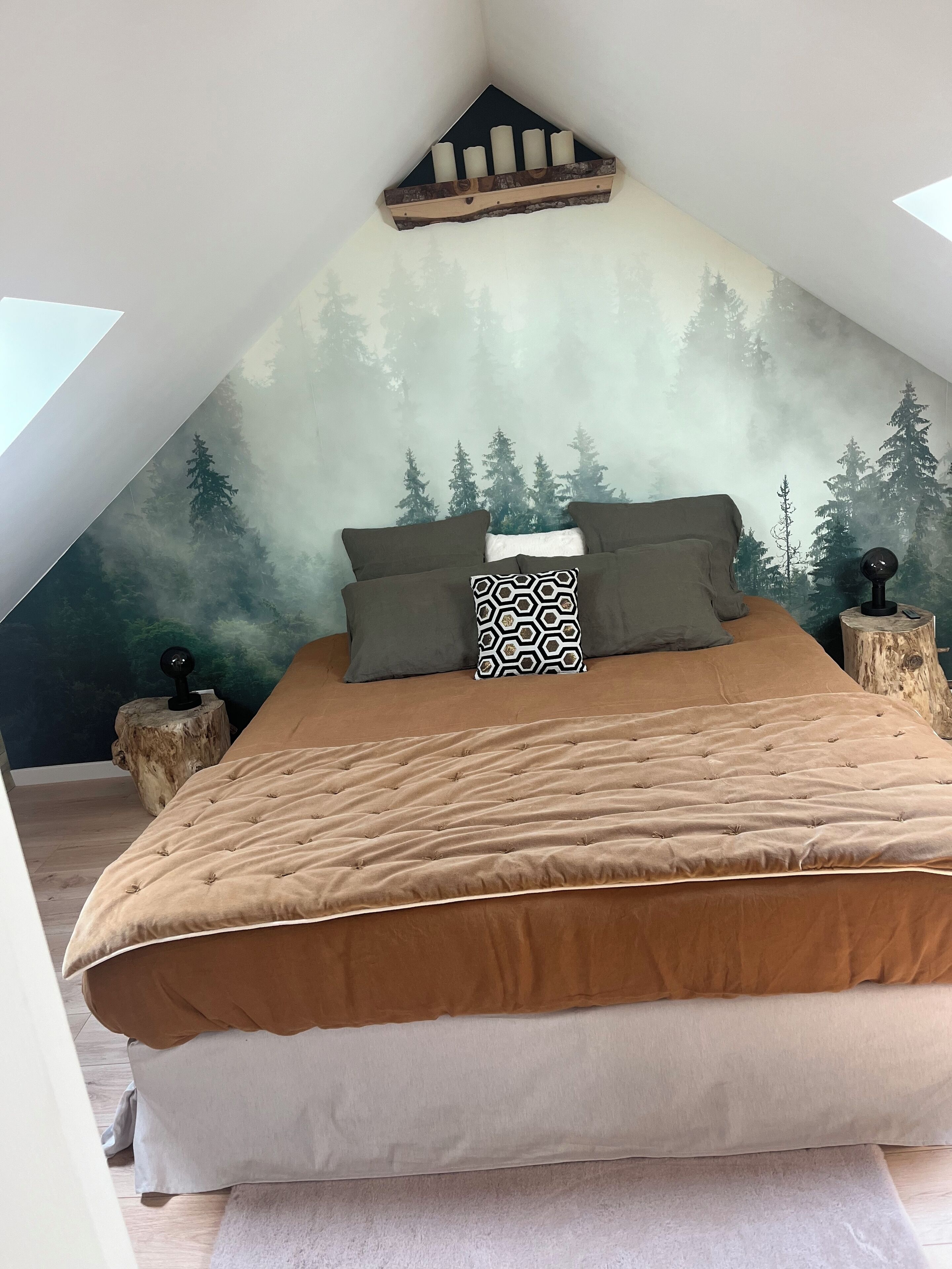A bedroom with a sloped ceiling, a bed with a brown bedspread, two pillows, and a wall mural of a forest.