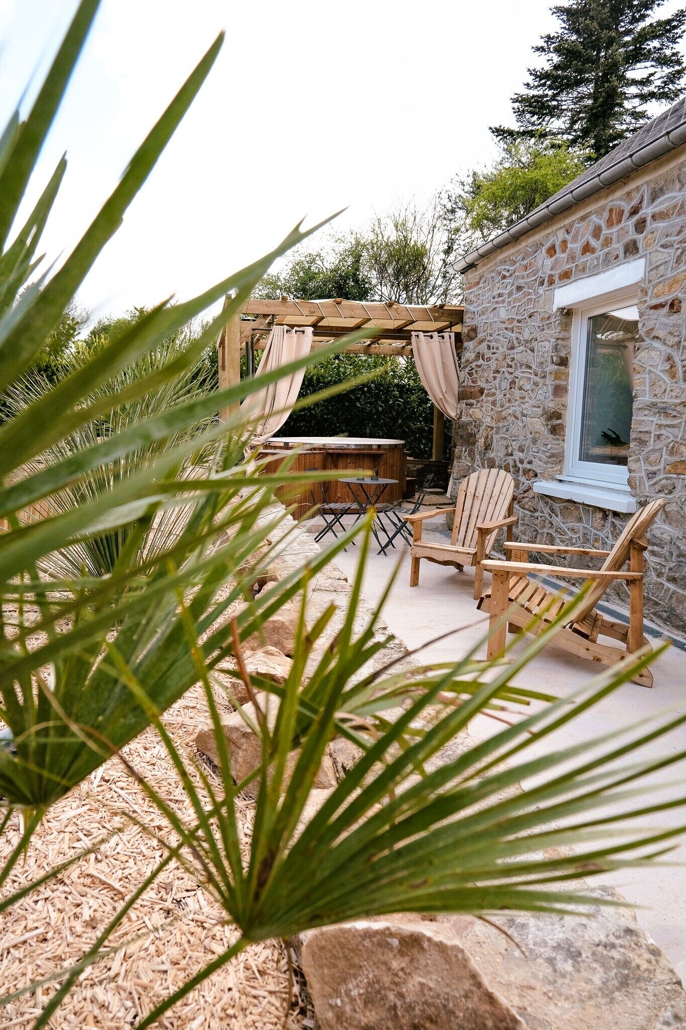 Ein Steinhaus mit einer Holzpergola, Außenbestuhlung und einem Fenster mit Blick auf einen Garten.