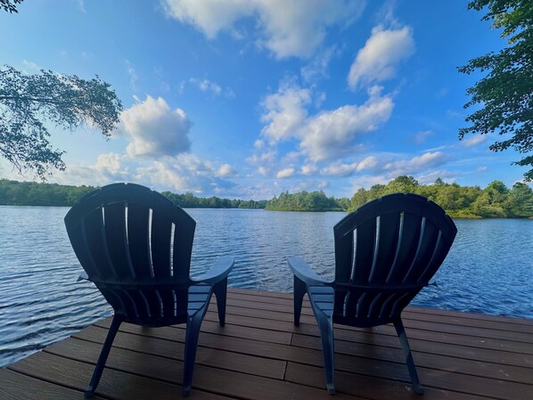 Two Adirondack chairs facing a lake with a wooden deck and a forested shoreline. - Lakefront House with Water Toys : close to Camelback, Kalahari, Casino, Hiking! (Long Pond)