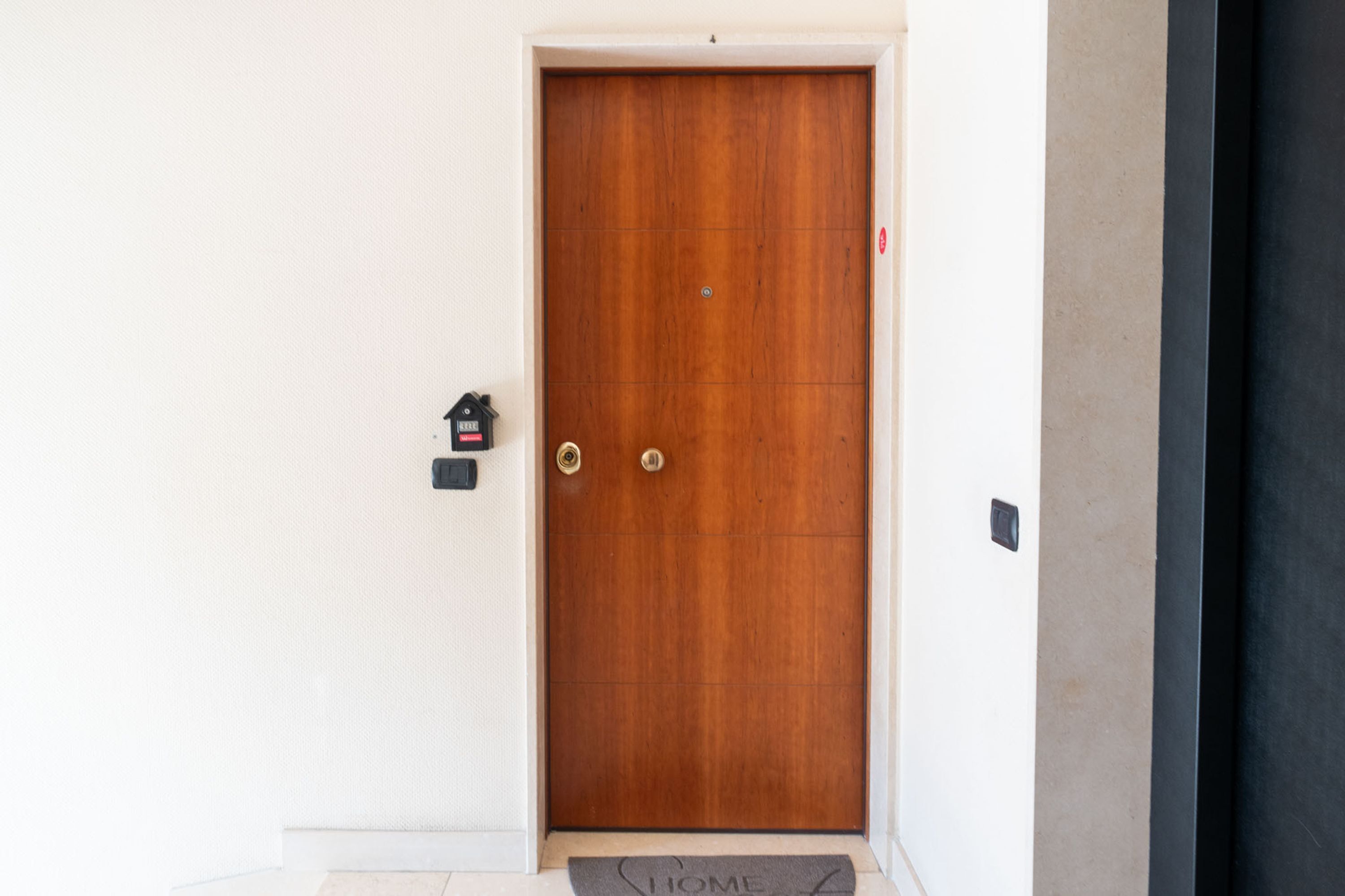 A wooden door with two handles, a doormat with "HOME" written on it, and a small mailbox on the wall to the left.
