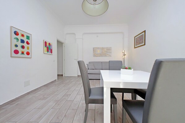 A modern dining area with a white table and chairs, a grey sofa, and two abstract paintings on the walls. - 4BNB - Cavalleggeri Apartment (Rome)