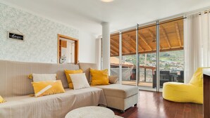 A living room with a sofa, yellow cushions, a round table, and a view through large glass doors. - Alfa I in Machico With 2 Bedrooms and 2 Bathrooms (Machico)