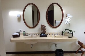 A bathroom with two sinks, a mirror, a towel dispenser, a soap dispenser, a potted plant, and a trash bin. - Natura Villa Ubud Bali (Bali)