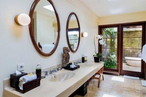 A bathroom with two sinks, two mirrors, a towel rack, a plant, and a bathtub visible through a glass door. - Natura Villa Ubud Bali (Bali)