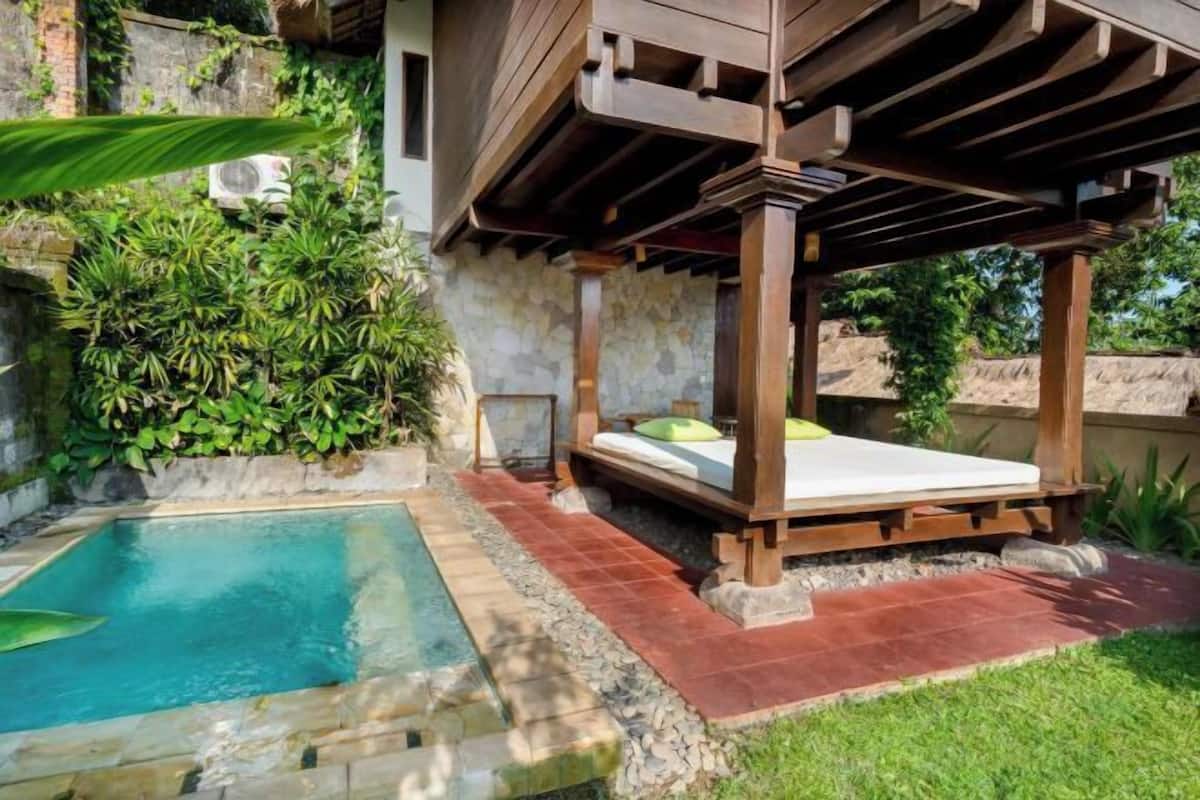 Un área junto a la piscina con una pérgola de madera, una cama pequeña con colchón y almohadas, y abundante vegetación.