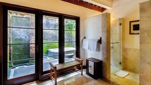 A bathroom with a glass shower, a bench, and a view of a garden through large windows. - Natura Villa Ubud Bali (Bali)