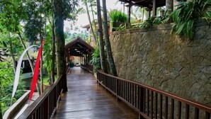 A wooden walkway with a stone wall on one side and a metal railing on the other, surrounded by greenery. - Natura Villa Ubud Bali (Bali)