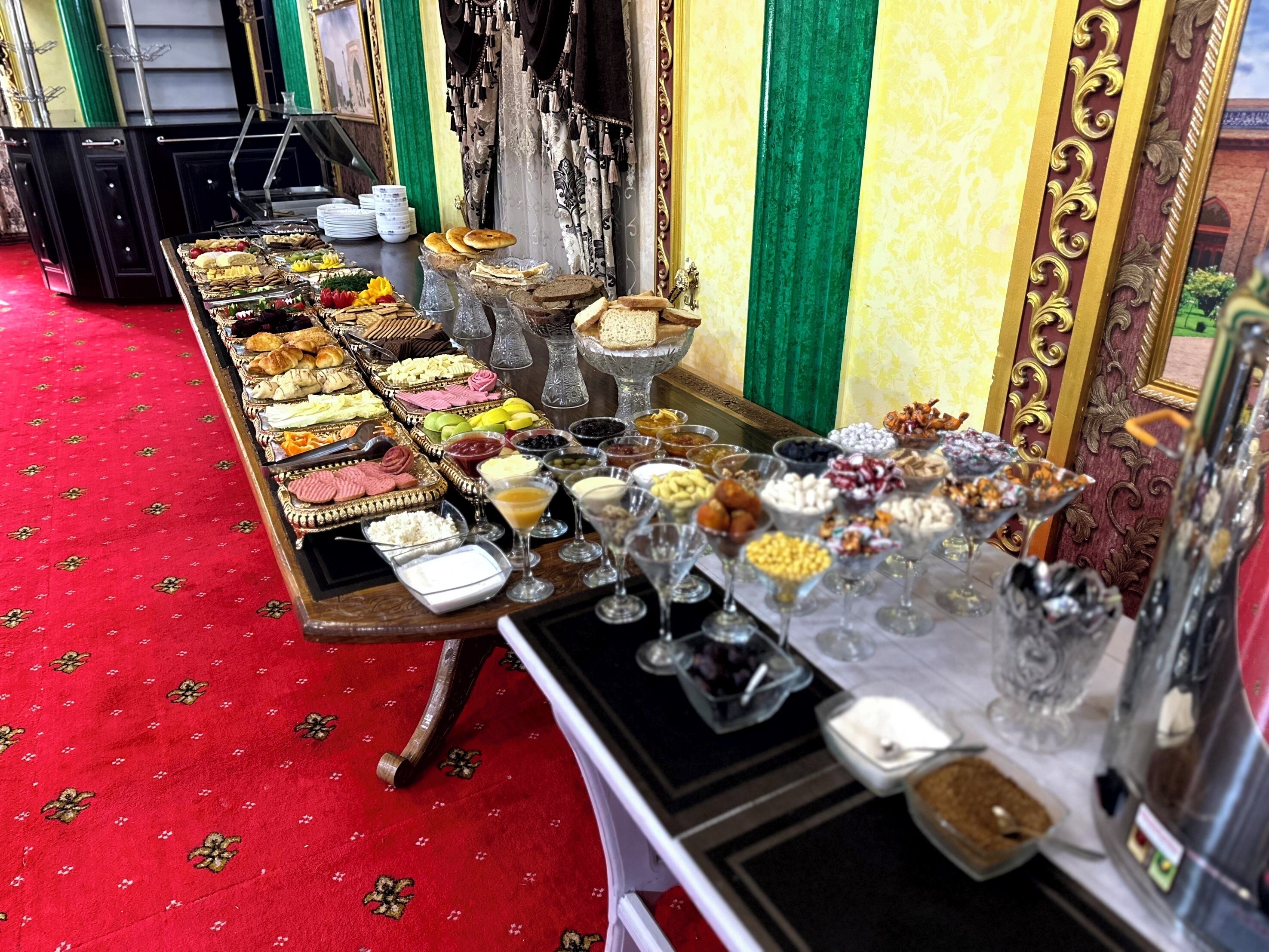 Una mesa de buffet con diversos alimentos y bebidas, sobre una alfombra roja con decoraciones ornamentales en la pared.