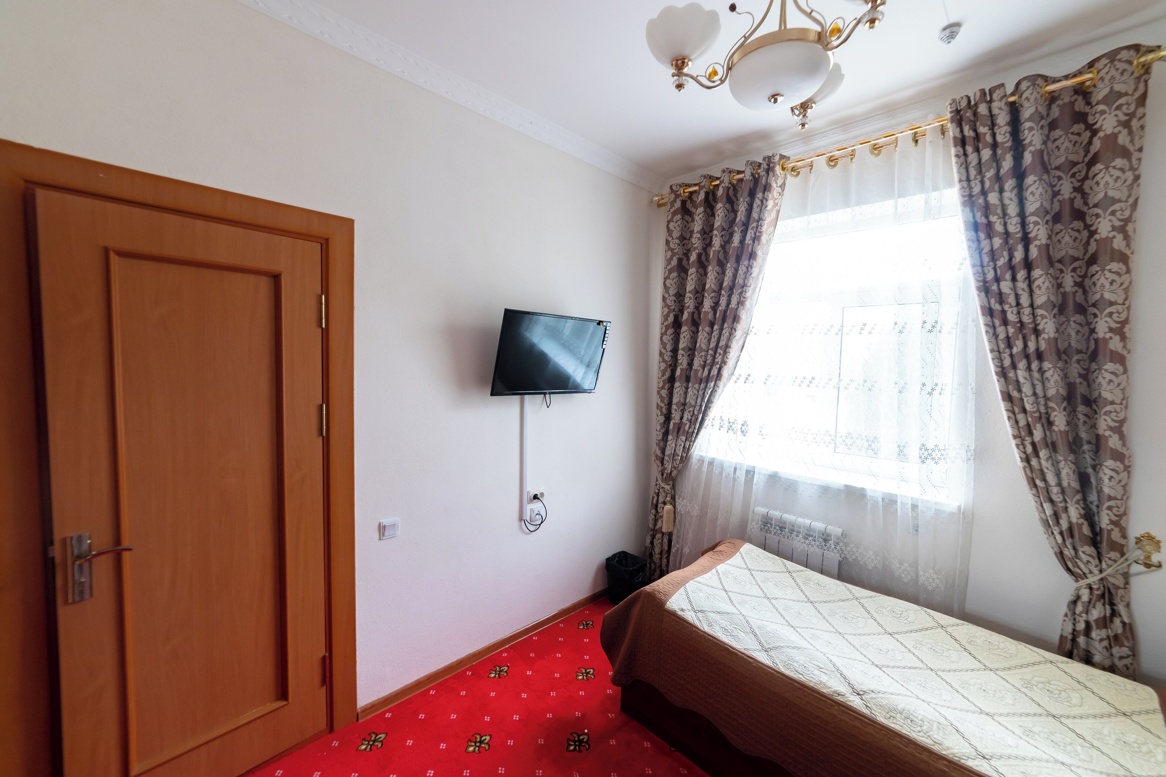 Een slaapkamer met een houten deur, een bed met een geborduurd beddegoed, een wandtelevisie en een raam met patroon gordijnen.