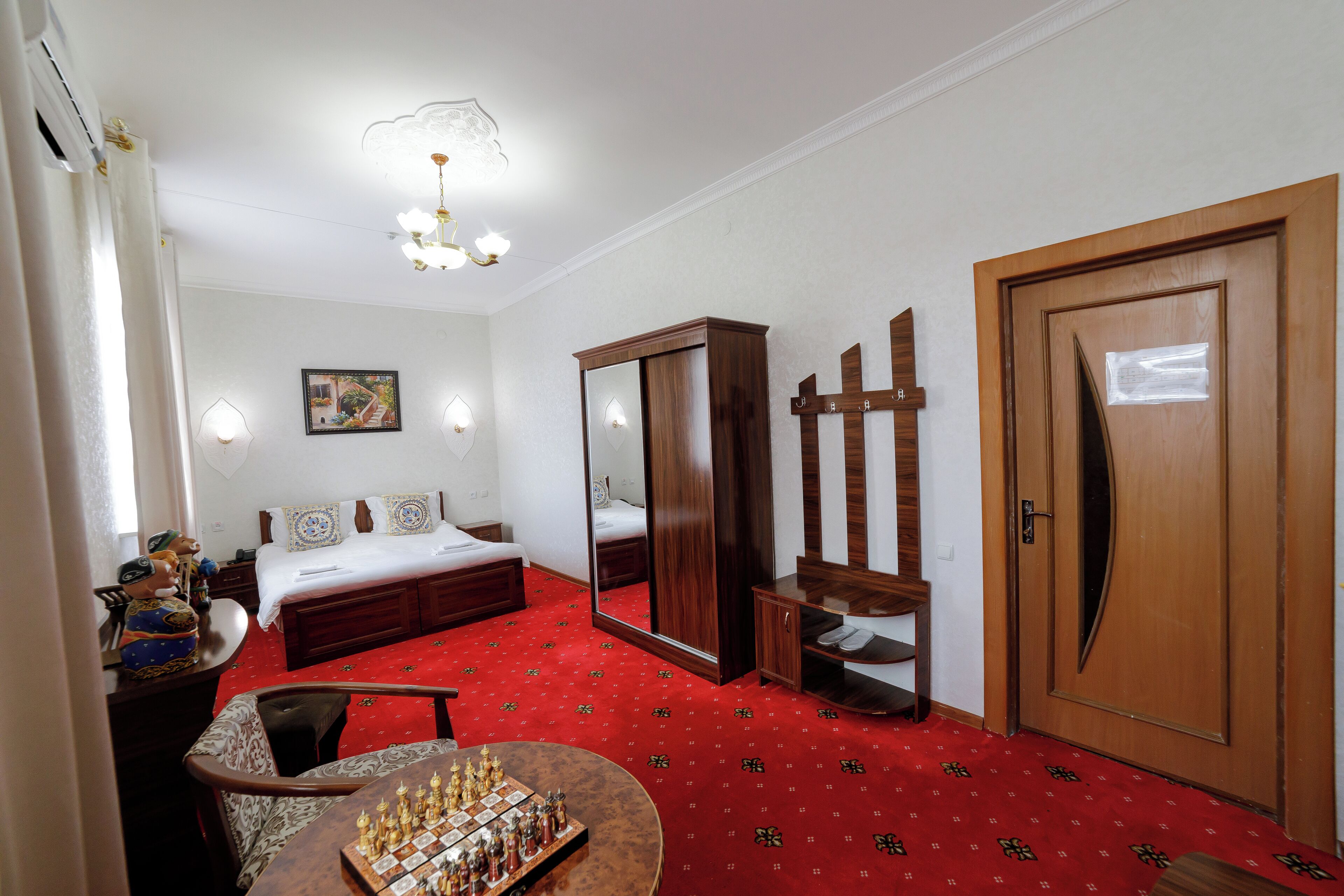 A hotel room with a red carpet, a wooden wardrobe, a large mirror, a bed with white bedding, and a chessboard on a table.