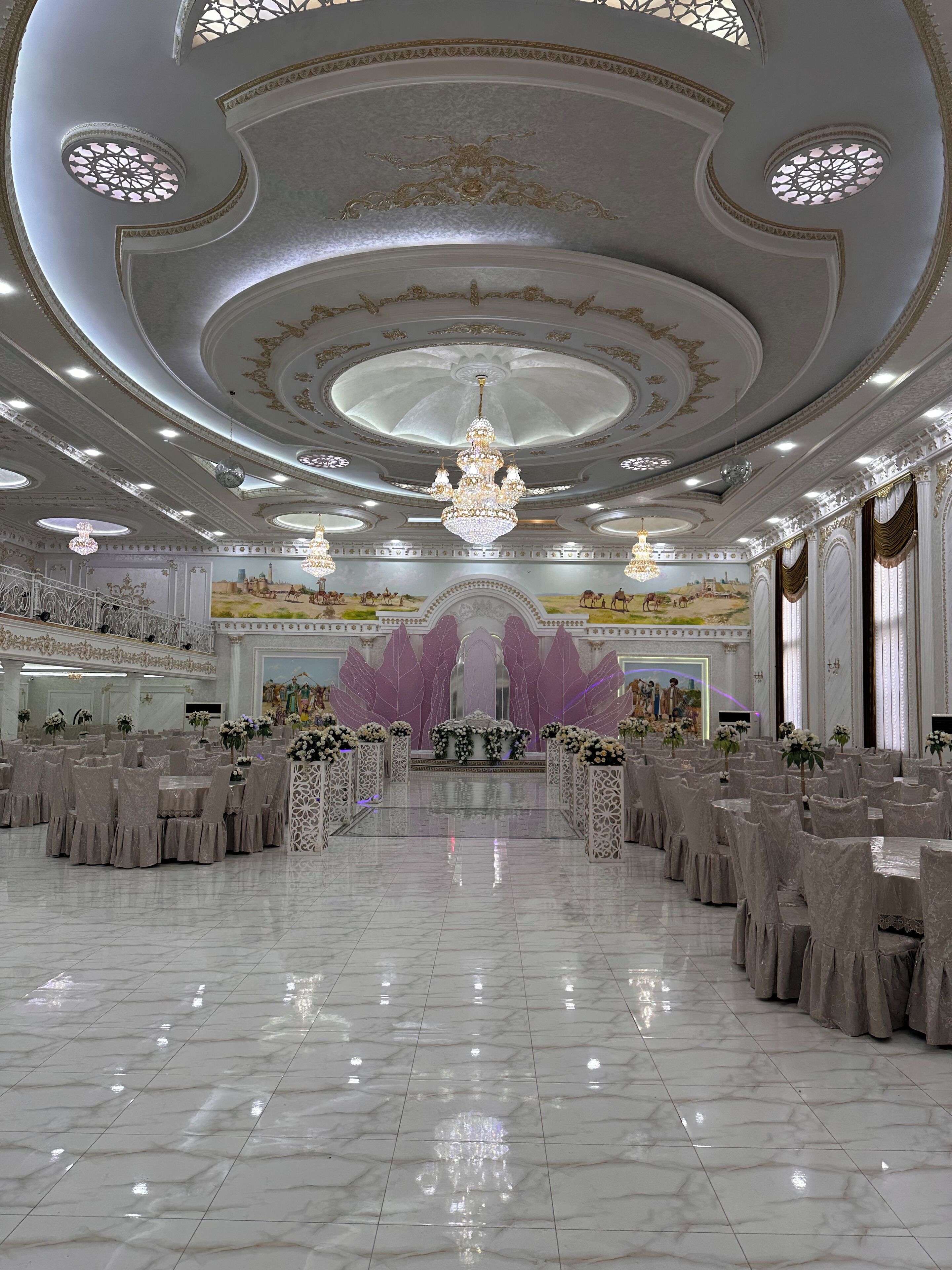 Un gran salón con candelabros ornamentales, un escenario central y un mural de gran tamaño en la pared de fondo.