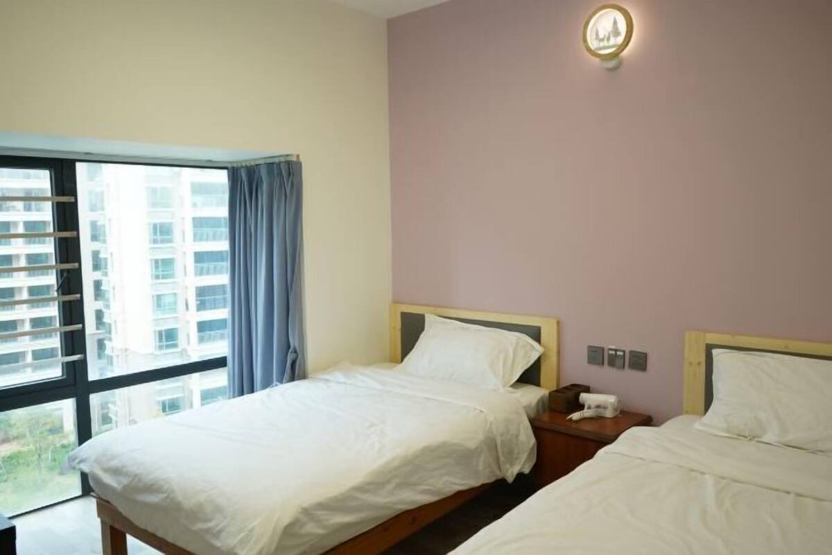 A hotel room with two beds, a window with curtains, a wall-mounted light, and a view of buildings outside.