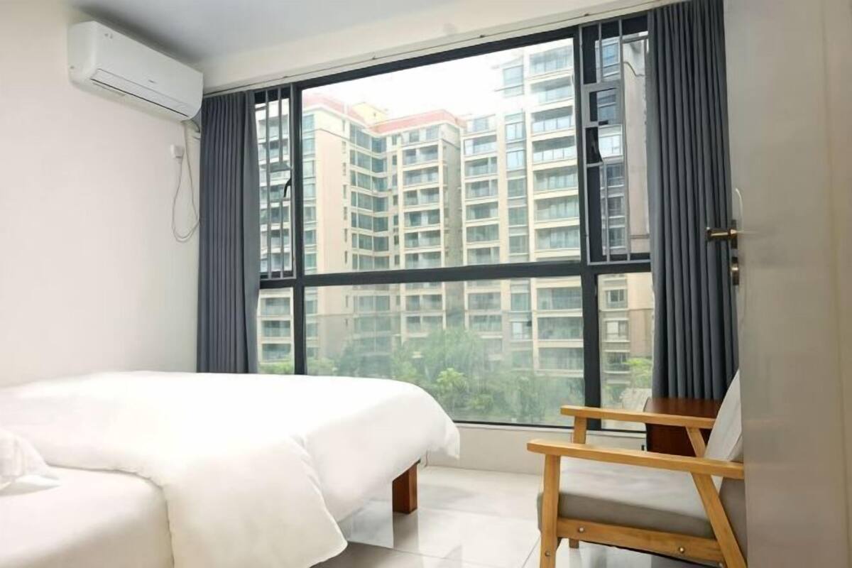 A hotel room with a bed, a small wooden table, a window with a view of high-rise buildings, and an air conditioning unit.