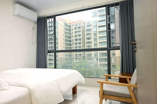 A hotel room with a bed, a small wooden table, a window with a view of high-rise buildings, and an air conditioning unit. - Youzhan Hostel (Shenzhen)