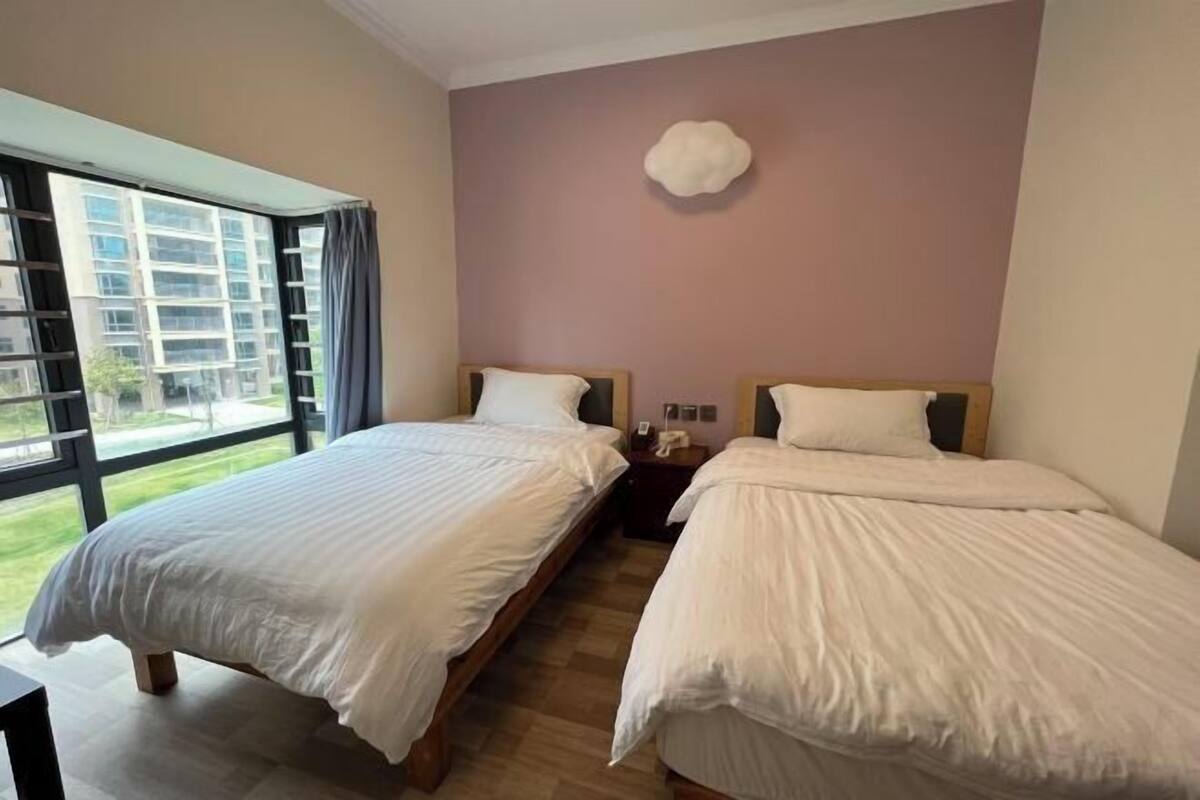 A hotel room with two beds, a large window with curtains, a wall-mounted light fixture, and a view of buildings outside.