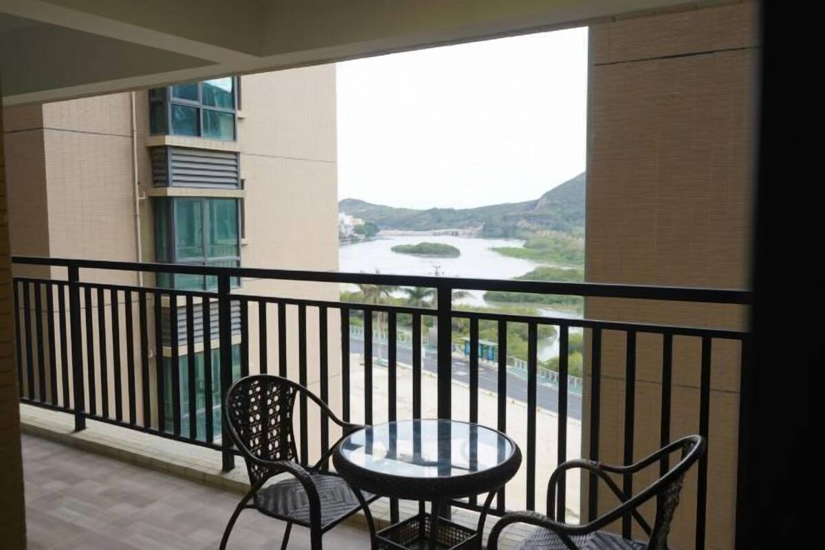 A balcony with a glass railing, two wicker chairs, and a small round table. A river and hills are visible in the background.