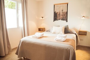 Una cama bien hecha con toallas, un tapiz en la pared y dos lámparas de noche en una habitación luminosa con un ventanal y cortinas.