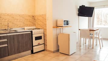 Een kleine keuken met een kookplaat, oven, magnetron en koelkast. Er is een eethoek met een tafel en stoelen.