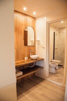 Un baño moderno con un mueble de tocador de madera, un espejo grande, un lavamanos, un inodoro y una zona de ducha.