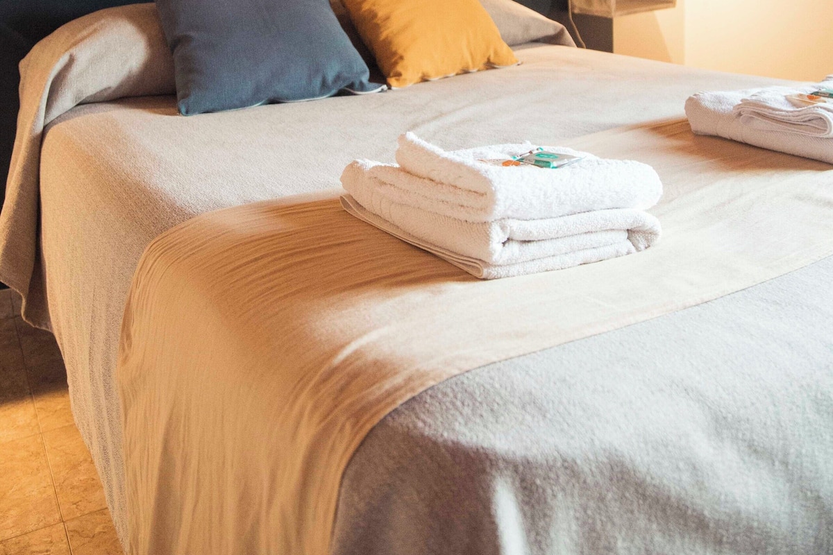 Una cama bien hecha con cabecera, almohadas y toallas dobladas en una habitación limpia y bien iluminada con techo de madera.