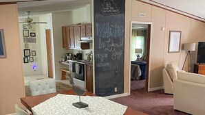 Un salon chaleureux avec une table à manger en bois, des chaises rembourrées de couleur gris clair, une petite table d’appoint et un tableau noir sur lequel est écrit un message de bienvenue.