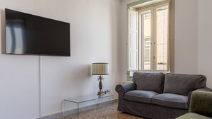 Een woonkamer met een grijze bank, een glazen salontafel met een lamp, een flatscreen televisie aan de muur en een raam met zonwering.