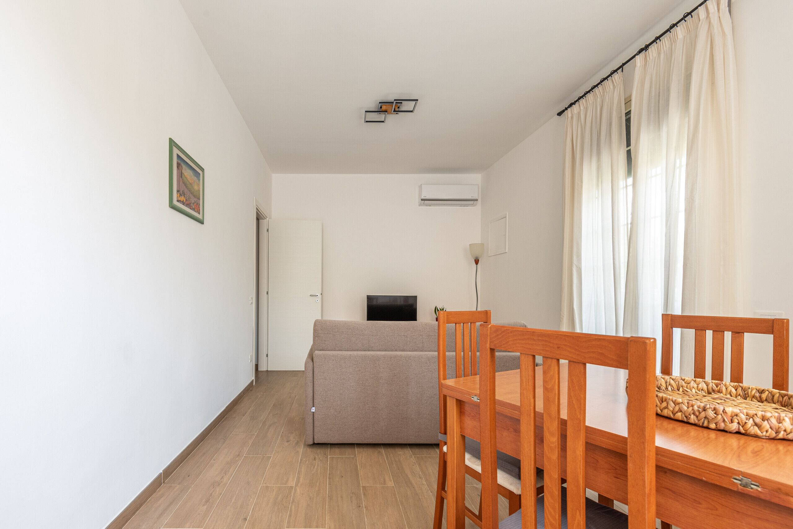 A room with a wooden dining table, a beige sofa, and a television on a stand.