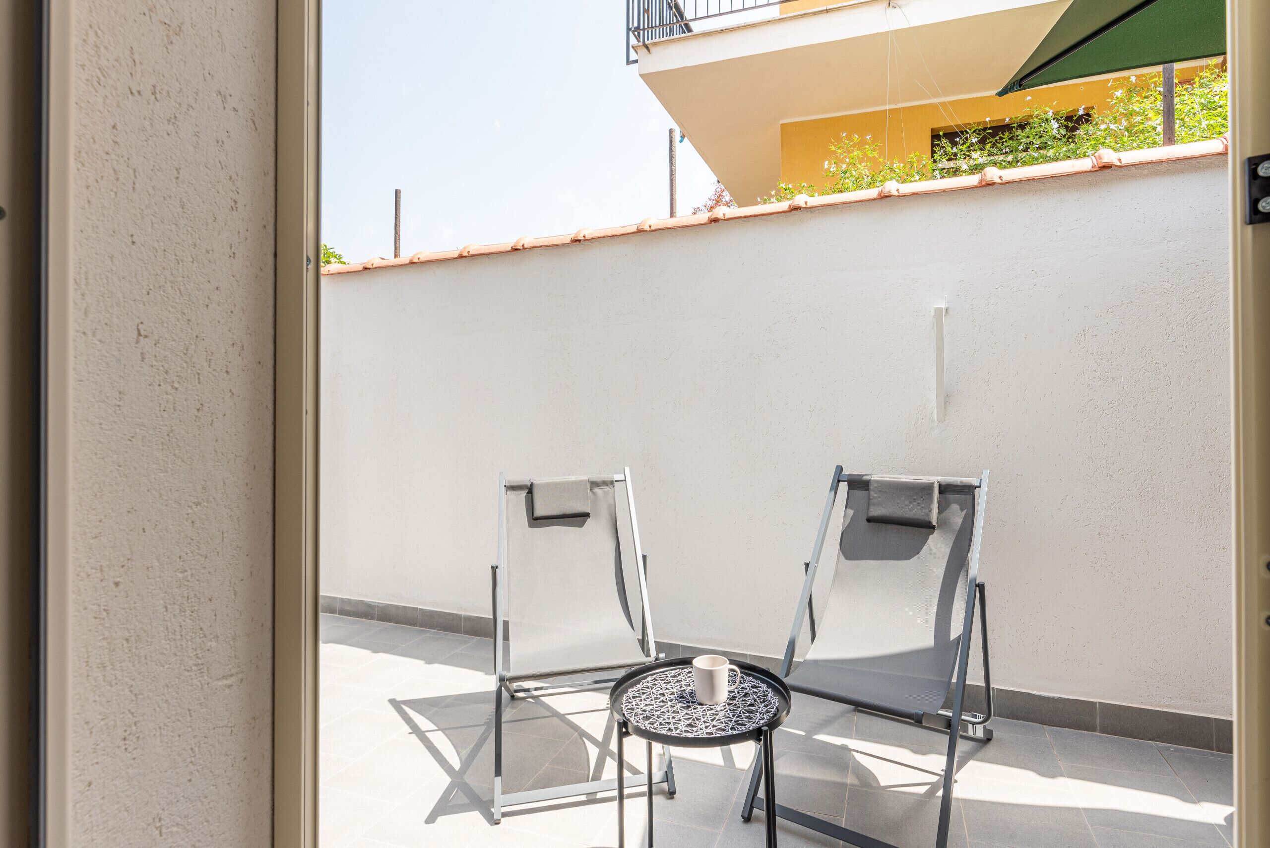 A balcony with two chairs and a small table, a white wall, and a tiled floor.