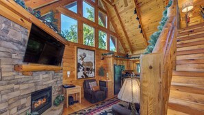A log cabin interior with a stone fireplace, wooden walls, and large windows. - Relaxing 3BD Cabin w/ HOT TUB! Pool Table + GAMES! (Sevierville)