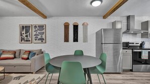 Una cocina moderna con un refrigerador de acero inoxidable, un horno empotrado y un microondas. Hay una zona de comedor con una mesa redonda y cuatro sillas. La cocina tiene paredes de ladrillo blanco y vigas de madera en el techo.