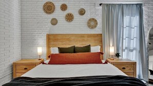 Habitación de hotel con pared de ladrillo blanco, cabecera de madera, cama con ropa de cama blanca y roja, dos mesitas de noche con lámparas y una ventana con cortinas.