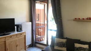 A small living area with a television, a coffee machine, a wooden cabinet, a sofa with cushions, and a sliding glass door leading to an outdoor balcony.