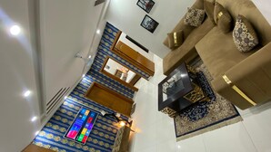 A living room with a sofa, a coffee table, and a TV. There is a patterned carpet and a blue and gold wall hanging. - Harvey's Apartment (Islamabad)