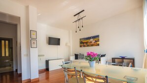 Un espace repas moderne avec une table en verre et des chaises en métal, un téléviseur à écran plat fixé au mur et un tableau représentant un paysage urbain.