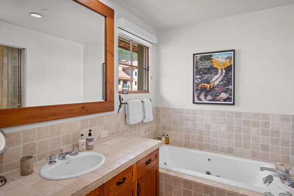 Een badkamer met een wastafel, spiegel en bad. Er is een raam met uitzicht op gebouwen en een lijst met een foto aan de muur.