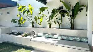 A modern outdoor seating area with concrete steps, a cushioned bench, and large potted plants against a white wall.