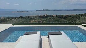 A pool with two lounge chairs facing a scenic view of the sea and hills.