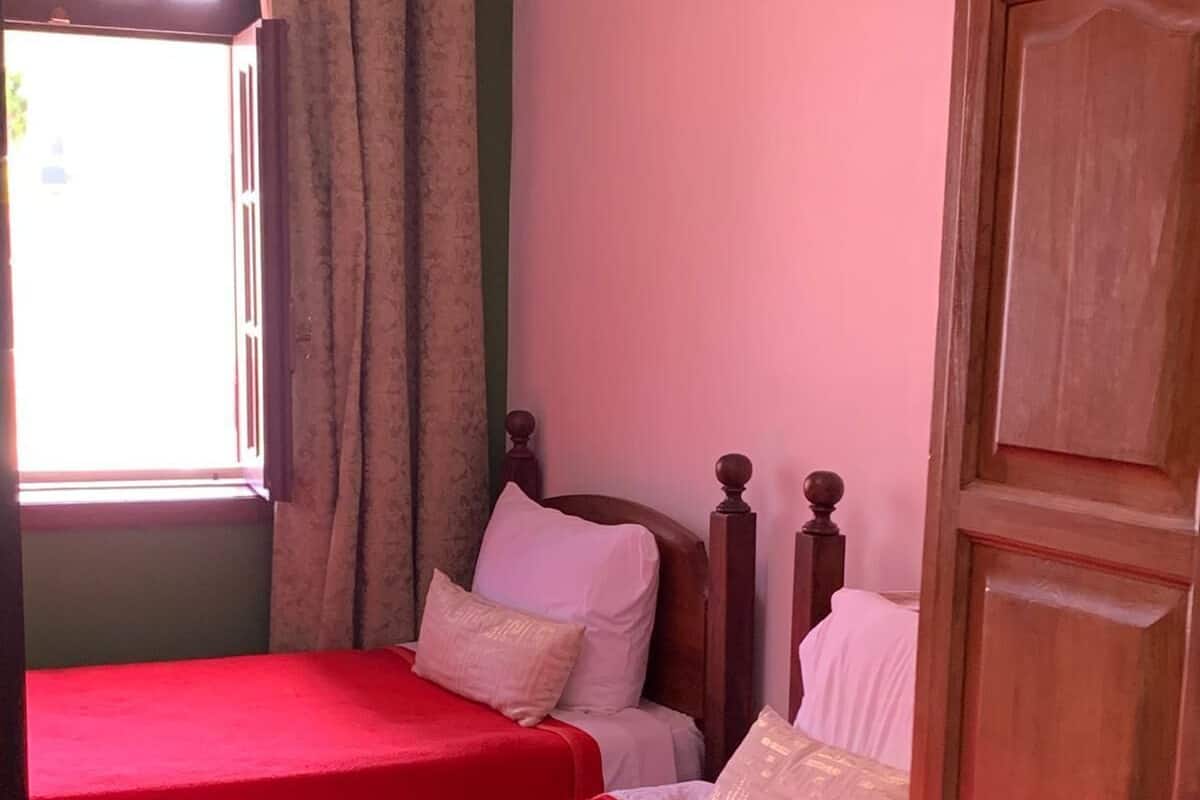 A room with two beds, each with a red bedspread, wooden headboards, and white bedding. A wooden wardrobe is visible on the right.