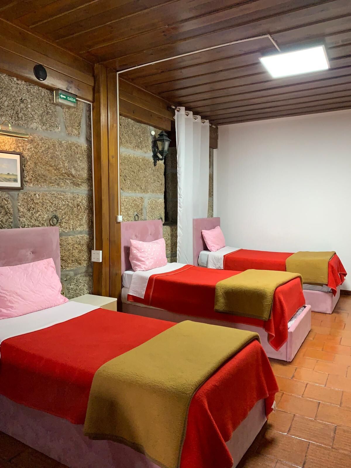 Quarto com três camas, cada uma com colcha vermelha e bege. Uma parede de pedra e teto de madeira.