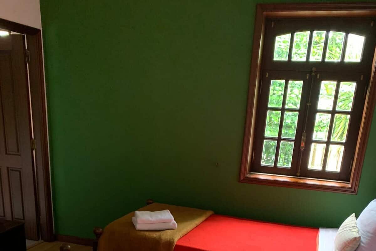 A room with two beds, each with a red bedspread and a brown headboard. There are towels on the beds, and a window with a view of greenery.