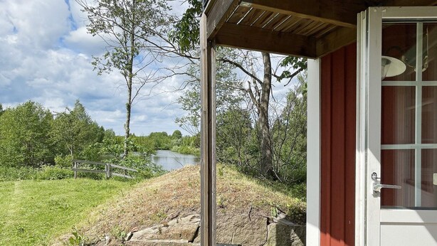Um deck de madeira com um muro de pedra, uma porta vermelha e vista para um lago e árvores.