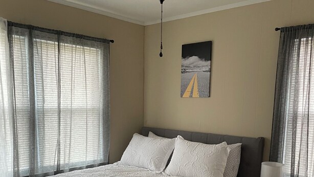 Et soveværelse med seng, ventilator, natbord med lampe og et billede på væggen.