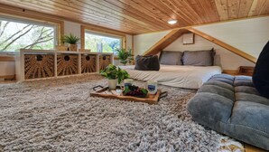 A cozy attic bedroom with a wooden ceiling, a large bed, a gray sofa, and a tray with fruits and mugs.