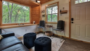 A cozy cabin interior with wooden walls, large windows offering forest views, a door with a keyhole, and a seating area with ottomans and chairs.