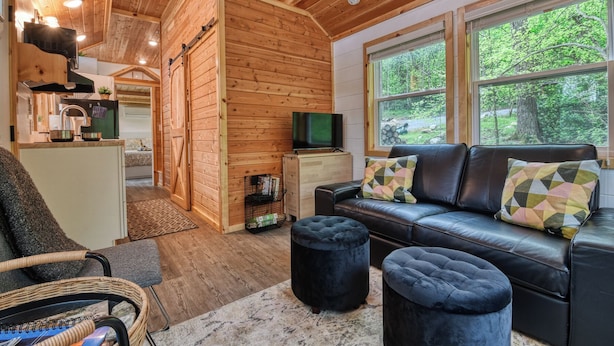 A cozy cabin interior with wooden walls, a sofa with patterned cushions, a television, and a view of the forest through large windows.