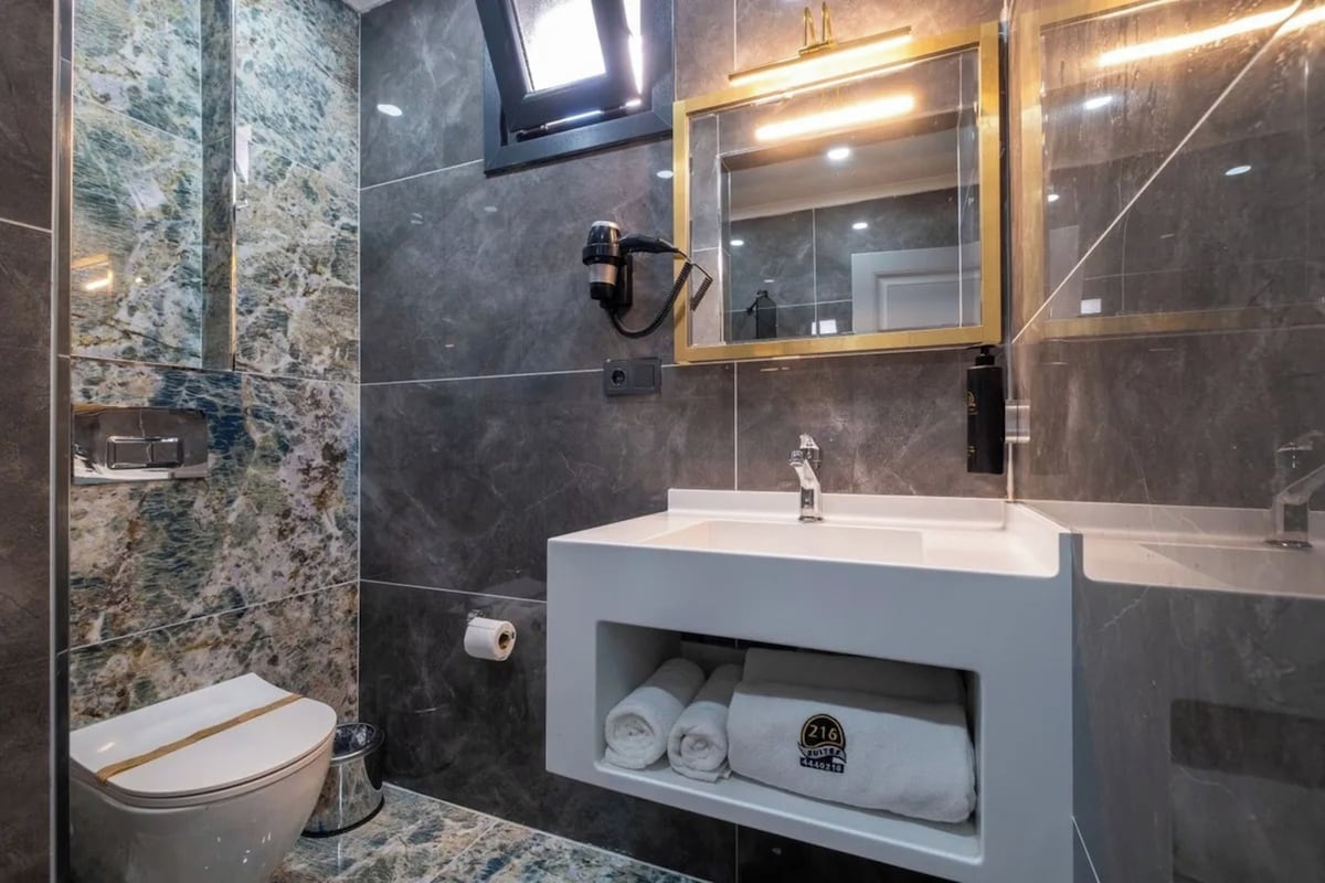 A modern bathroom with a marble backsplash, a white sink with a gold faucet, a mirror with a gold frame, and a towel shelf.