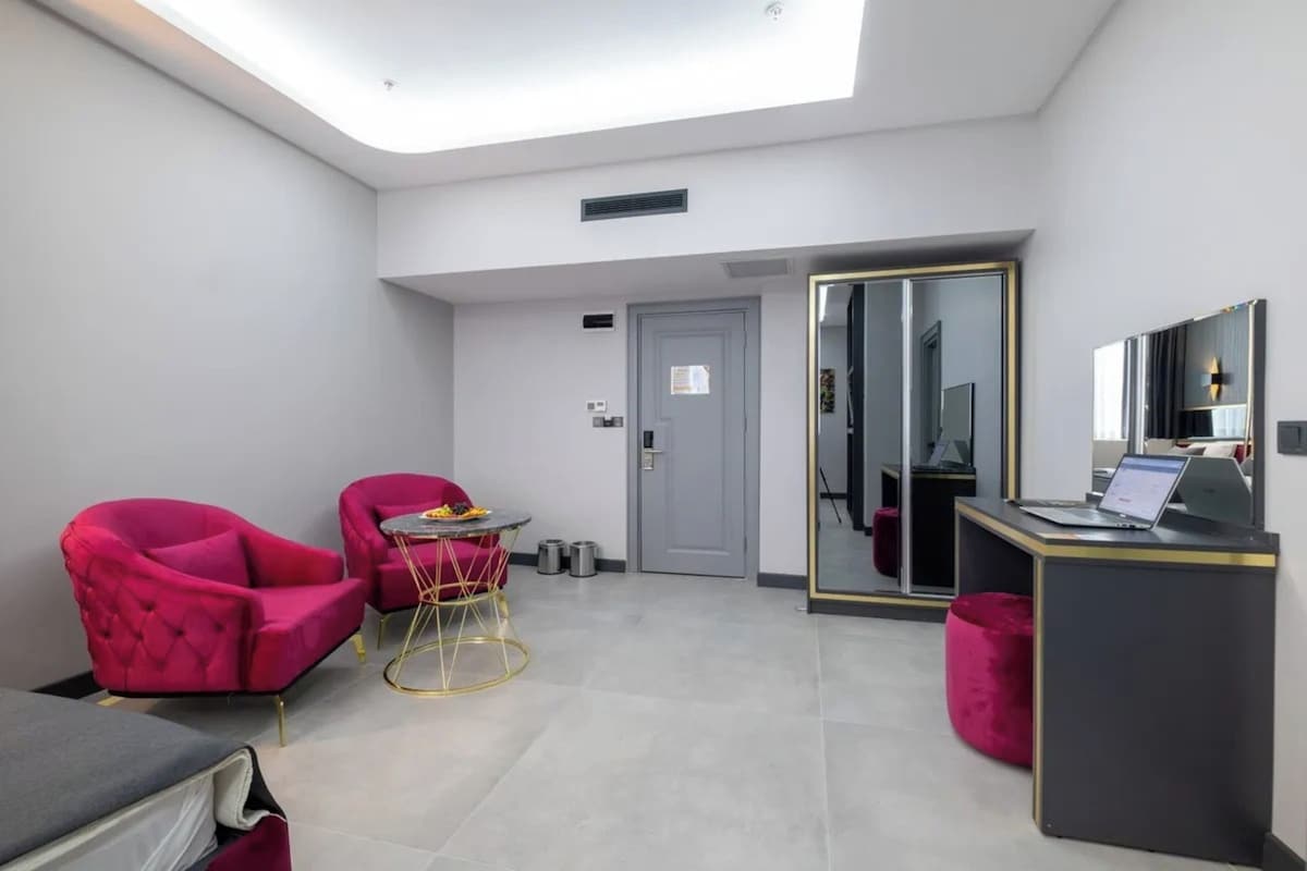 A modern hotel room with a skylight, a desk with a laptop, two pink armchairs, and a small round table.