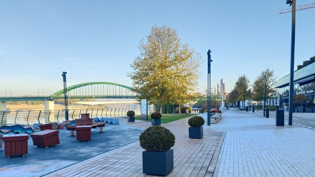 Un passeig marítim amb bancs de fusta, plantes en testos i un pont modern al fons.
